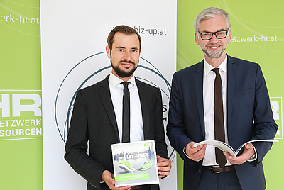 Zwei Männer stehen nebeneinander und halten eine Broschüre in der Hand. Der Blick ist zur Kamera gerichtet